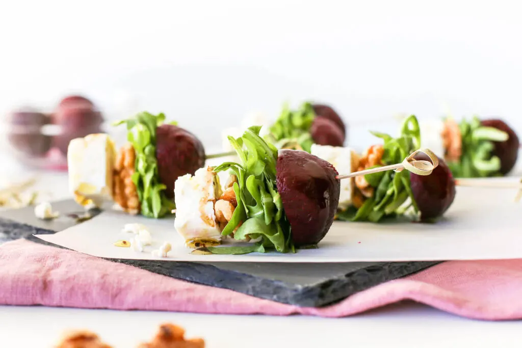 Goat cheese, walnuts, arugula, and roasted beets speared with a stick served atop a slate platter on a purple napkin.