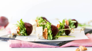Goat cheese, walnuts, arugula, and roasted beets speared with a stick served atop a slate platter on a purple napkin.