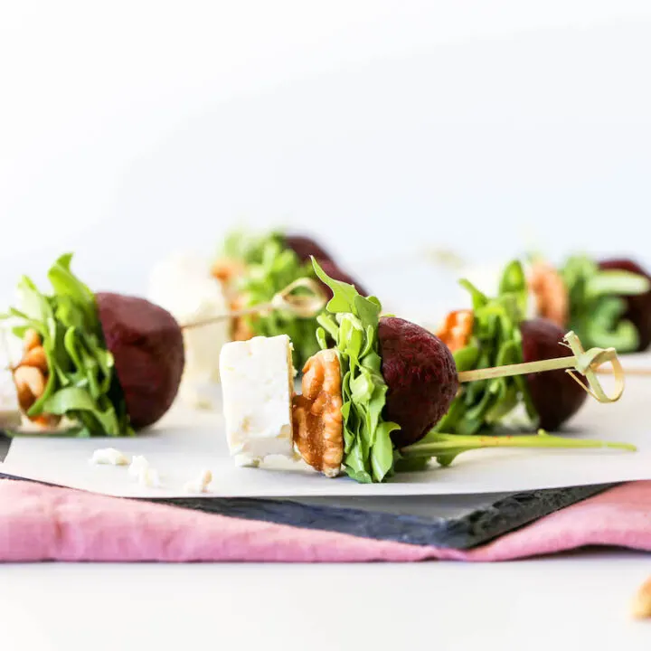 Goat cheese, walnuts, arugula, and roasted beets speared with a stick served atop a slate platter on a purple napkin.