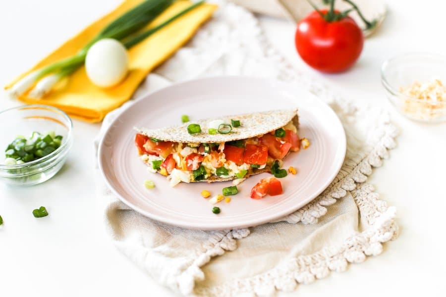 A hard boiled egg quesadilla on a pink plate atop a cream-colored towel with ingredients surrounding the plate including chopped green onions, a hard boiled egg, a whole tomato, and some cheese in a bowl