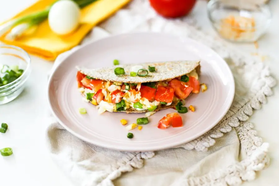 A hard boiled egg quesadilla on a pink plate atop a cream-colored towel with ingredients surrounding the plate including chopped green onions, a hard boiled egg, a whole tomato, and some cheese in a bowl