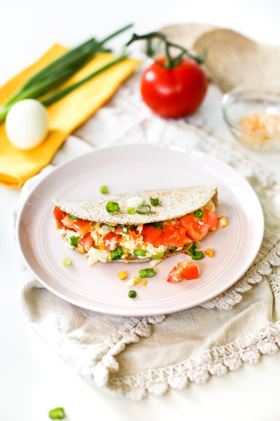 A hard boiled egg quesadilla on a pink plate atop a cream-colored towel with ingredients surrounding the plate including chopped green onions, a hard boiled egg, a whole tomato, and some cheese in a bowl