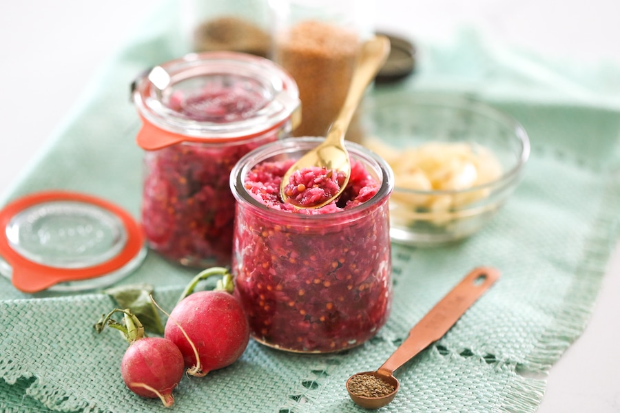 Two jars of radish relish on a blue placemat surrounded by ingredients