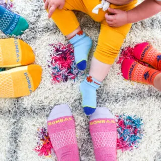 Bird's eye view of baby in yellow pants and blue Bombas socks surrounded by feet wearing colorful Bombas socks