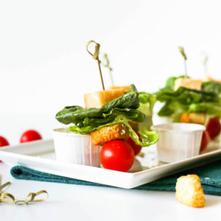 Tomato, crouton, romaine lettuce, and a cube of parmesan cheese speared by a toothpick, leaning against a white condiment cup on a serving platter.