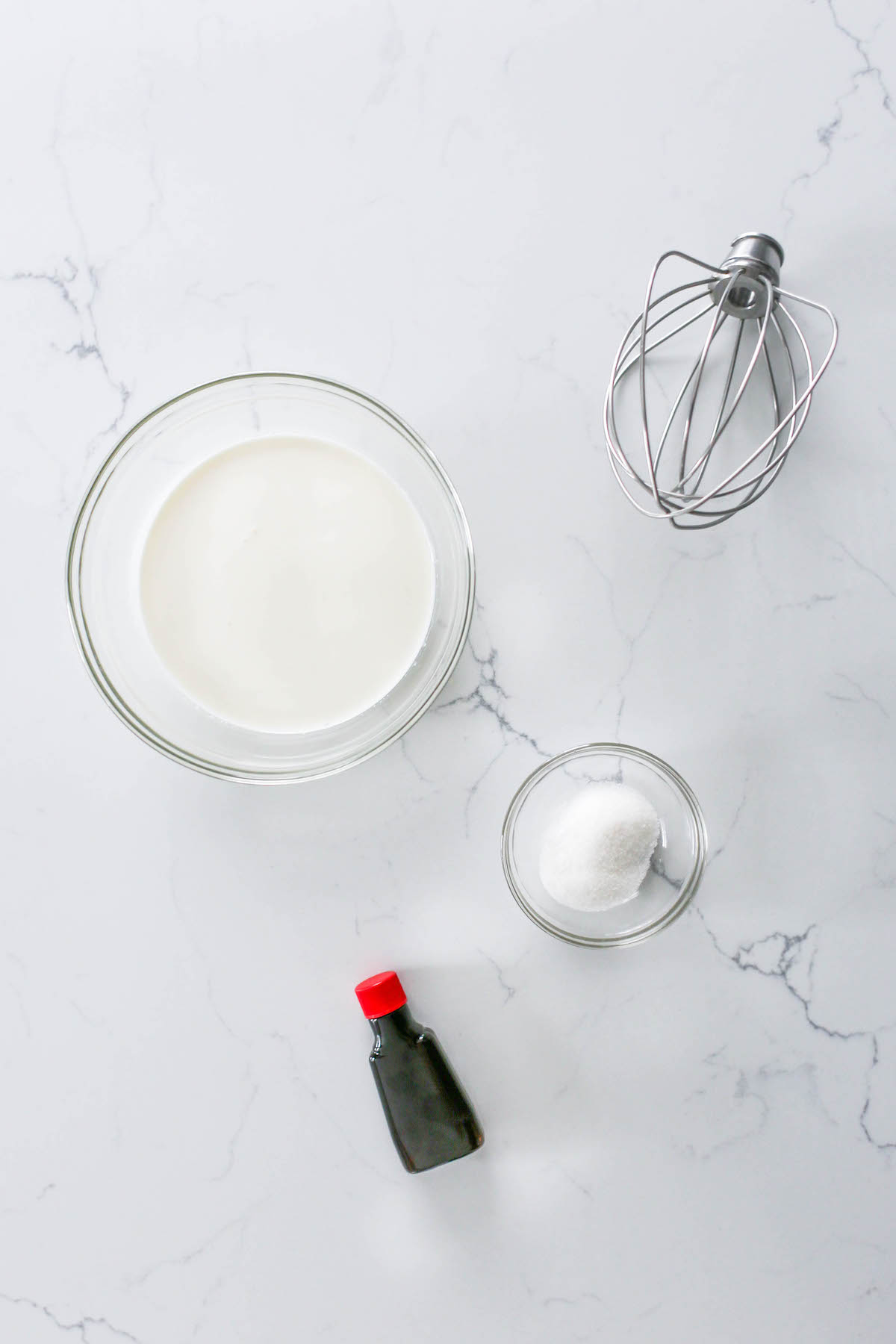 Overhead view of heavy whipping cream in a bowl, vanilla extract bottle, a small bowl of granulated sugar, and a KitchenAid Stand Mixer whisk attachment on a marble countertop.