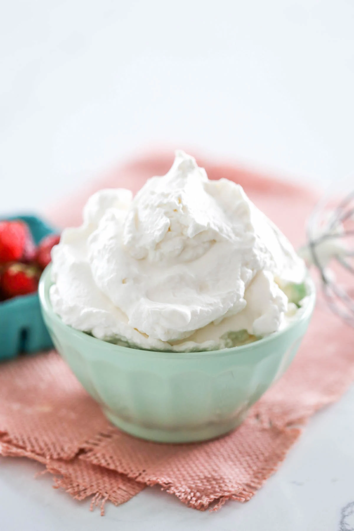 Homemade whipped cream in a mint colored bowl atop a pink placement with a carton of strawberries in the background