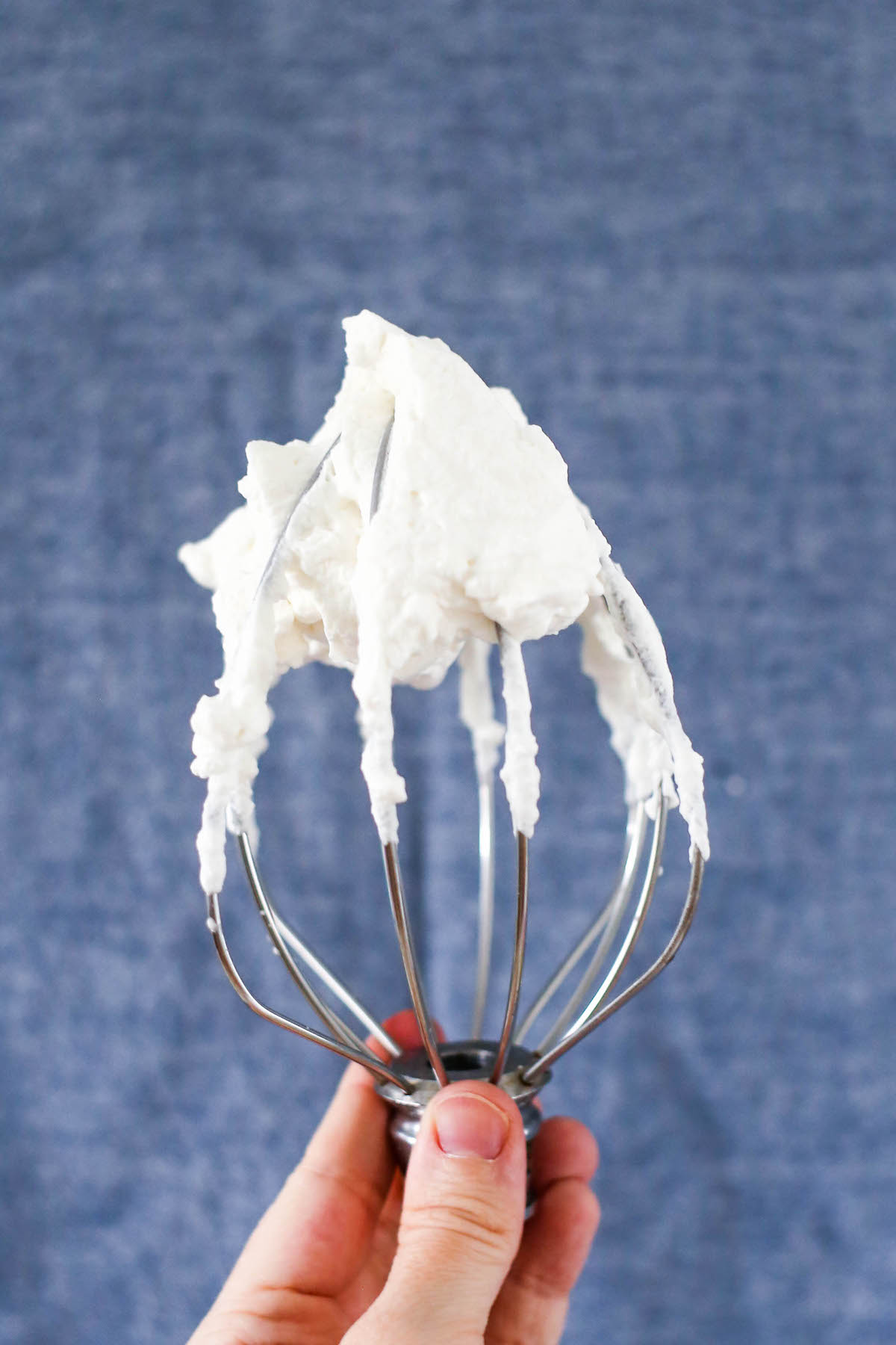 A KitchenAid mixer whisk attachment with whipped cream on it held in front of a dark blue background