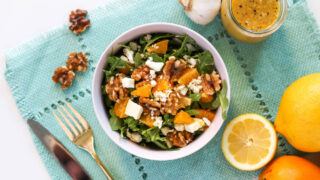 Overhead view of arugula, oranges, walnuts, and feta cheese in a pink salad bowl atop a blue placemat with gold flatware, walnuts, a slice of lemon, a head of garlic, and a jar of vinaigrette salad dressing off to the side