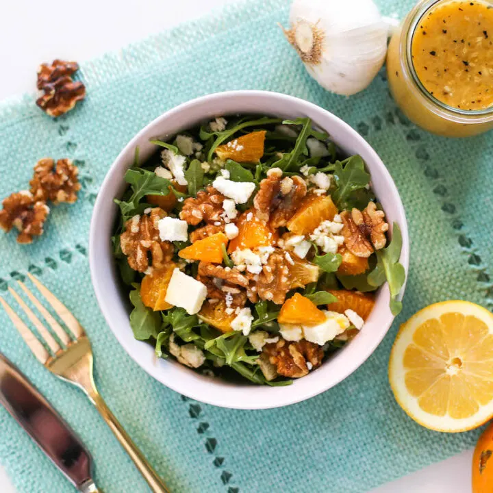 Overhead view of arugula, oranges, walnuts, and feta cheese in a pink salad bowl atop a blue placemat with gold flatware, walnuts, a slice of lemon, a head of garlic, and a jar of vinaigrette salad dressing off to the side