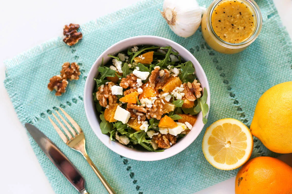 Overhead view of arugula, oranges, walnuts, and feta cheese in a pink salad bowl atop a blue placemat with gold flatware, walnuts, a slice of lemon, a head of garlic, and a jar of vinaigrette salad dressing off to the side