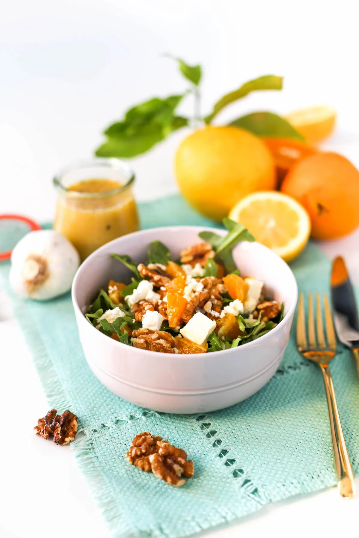 Orange and arugula salad in a pink bowl with walnuts and feta cheese atop a blue placemat with walnuts, gold flatware, sliced and whole lemons and oranges, a head of garlic, and a jar of lemon vinaigrette around it