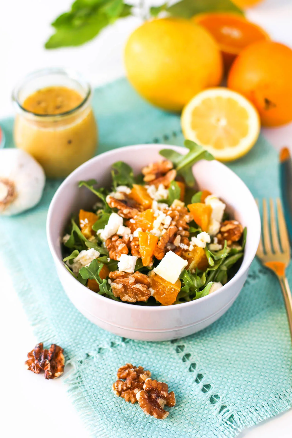 Orange and arugula salad in a pink bowl with walnuts and feta cheese atop a blue placemat with walnuts, gold flatware, sliced and whole lemons and oranges, a head of garlic, and a jar of lemon vinaigrette around it