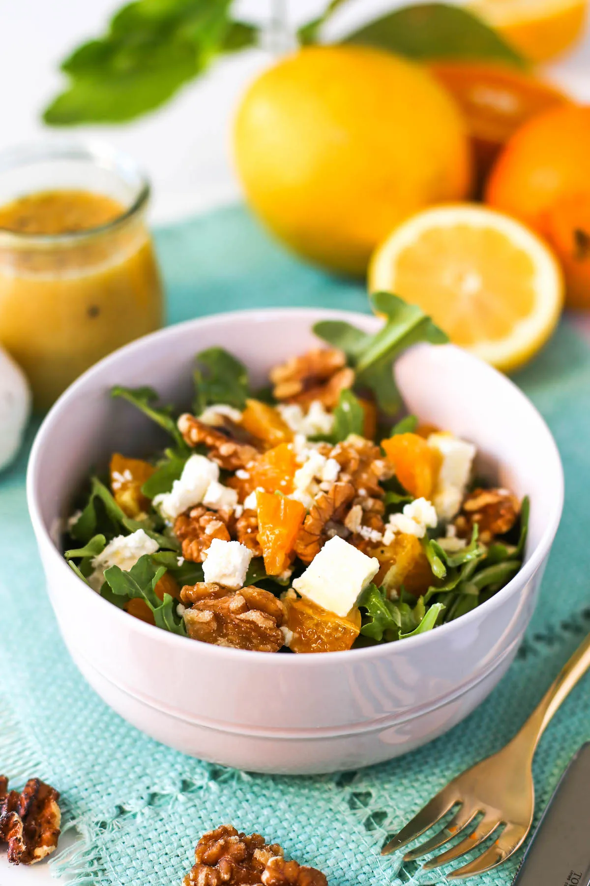 Orange and arugula salad in a pink bowl with walnuts and feta cheese atop a blue placemat with walnuts, gold flatware, sliced and whole lemons and oranges, a head of garlic, and a jar of lemon vinaigrette around it
