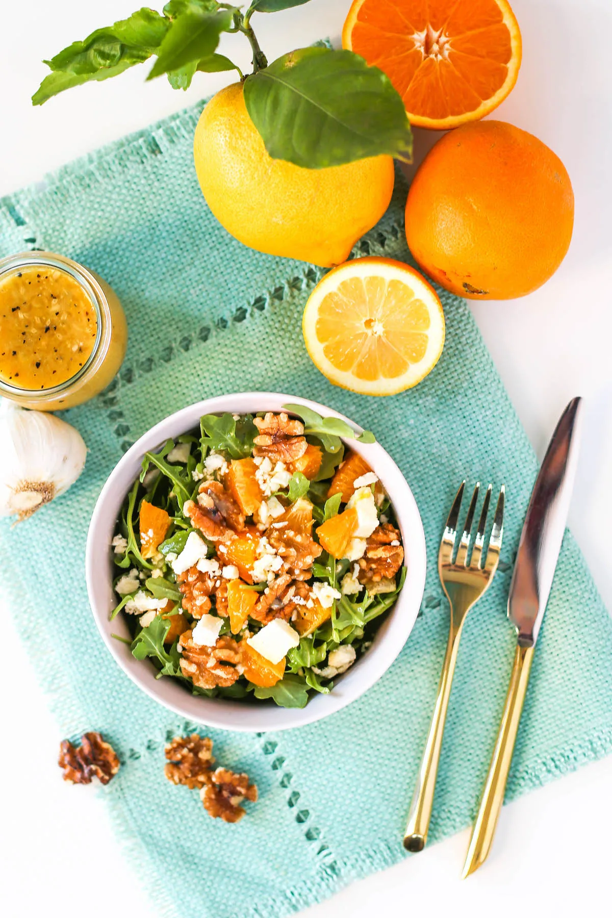 Overhead view of arugula, oranges, walnuts, and feta cheese in a pink salad bowl atop a blue placemat with gold flatware, walnuts, a slice of lemon, a head of garlic, and a jar of vinaigrette salad dressing off to the side