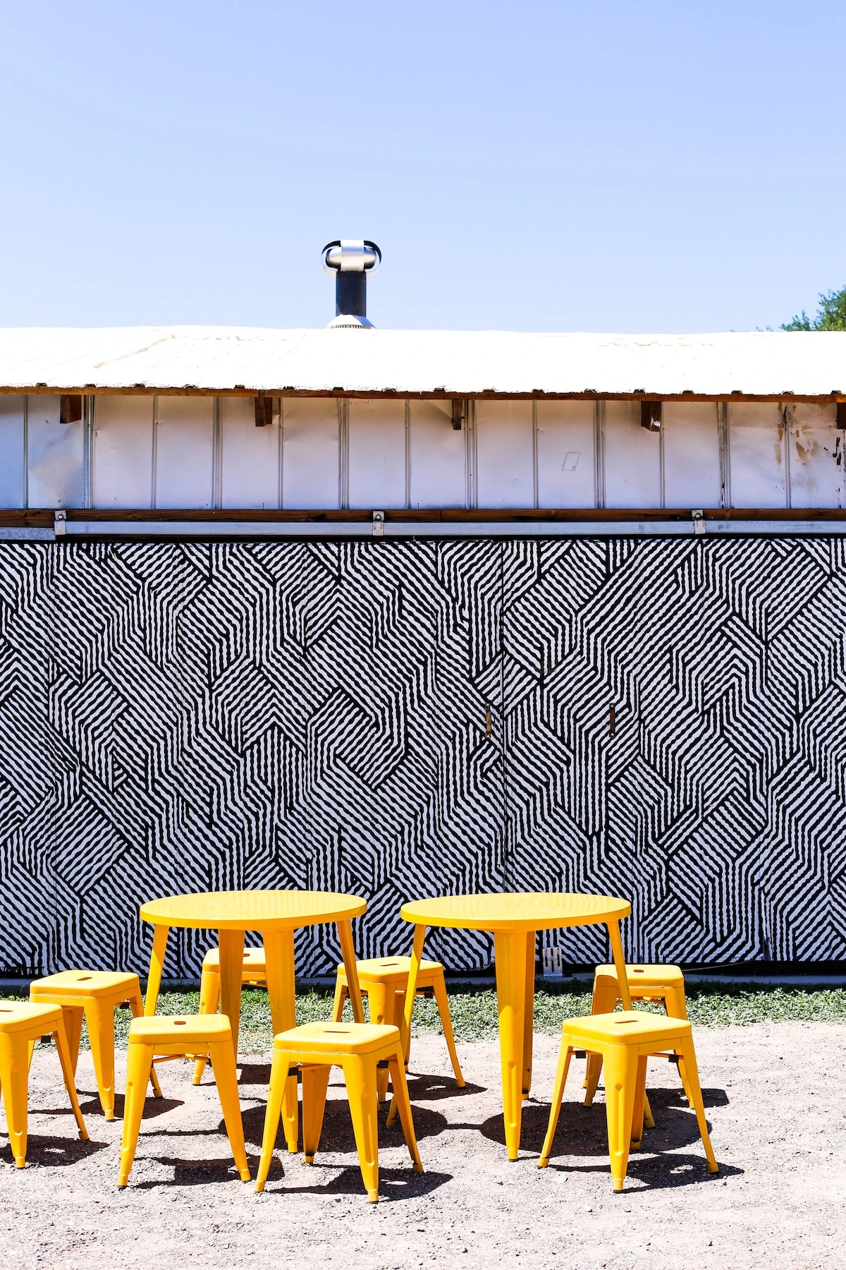 Yellow table and stools outside a building with a black and white geometric mural painted on the side.