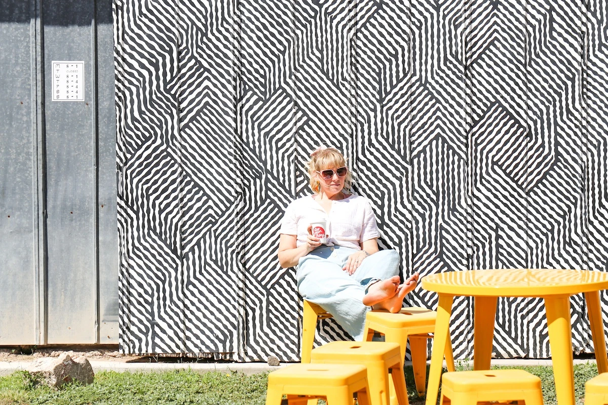 Woman holding a paper coffee cup while sitting on yellow stool at a yellow table in front of a building painted with a black and white geometric mural