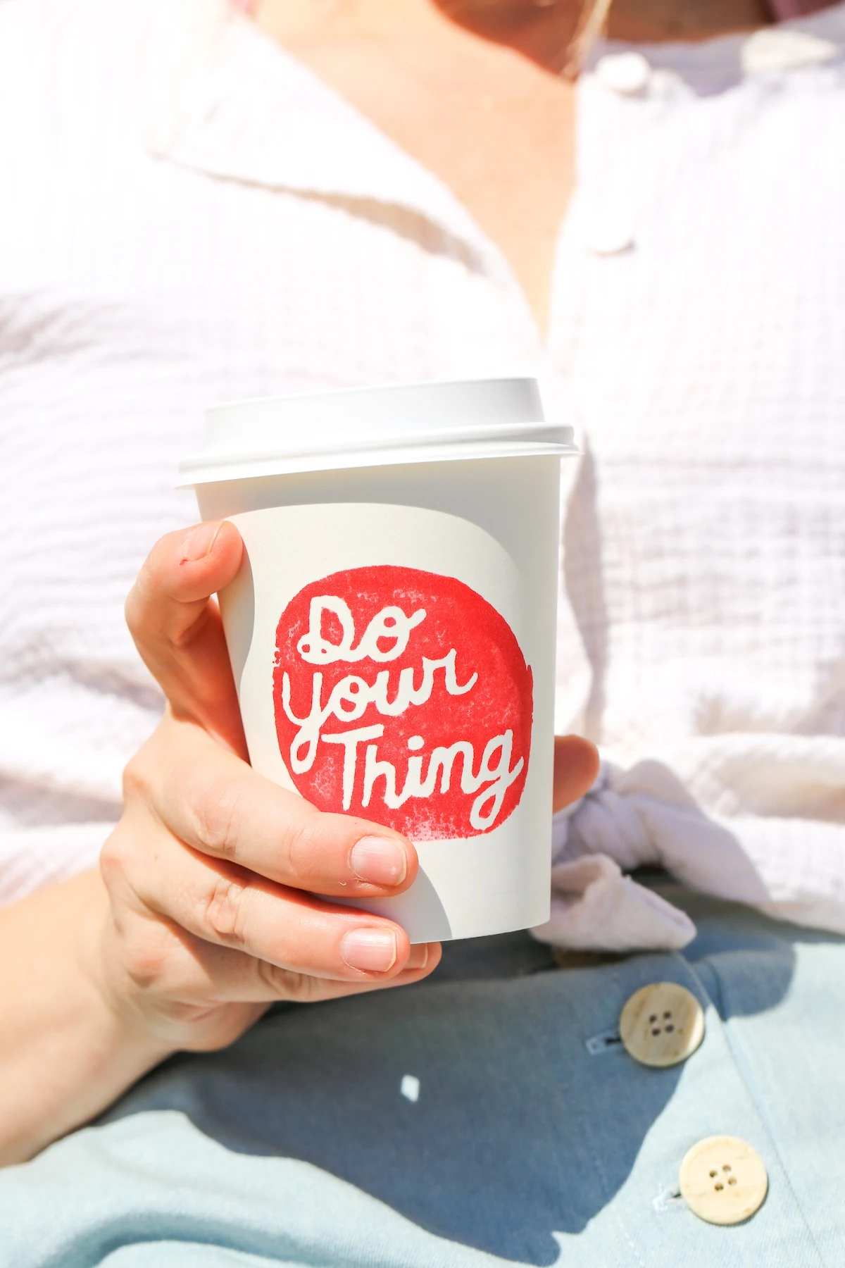 Close-up of woman's hand holding a white paper coffee up with the logo reading "Do Your Thing" on a white circle.