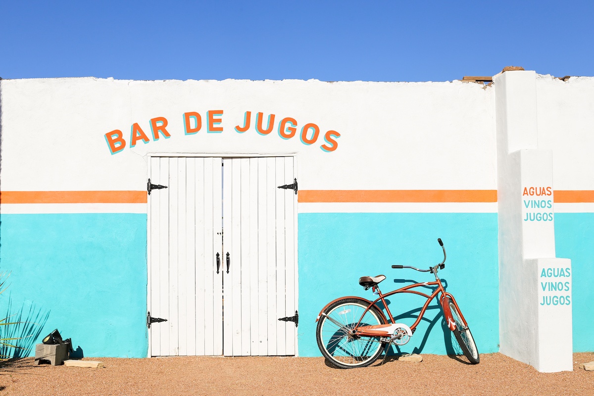 White and turquoise wall with orange stripe outside of restaurant with "Bar de Jugos" painted above white wooden doors and bike leaning against wall.