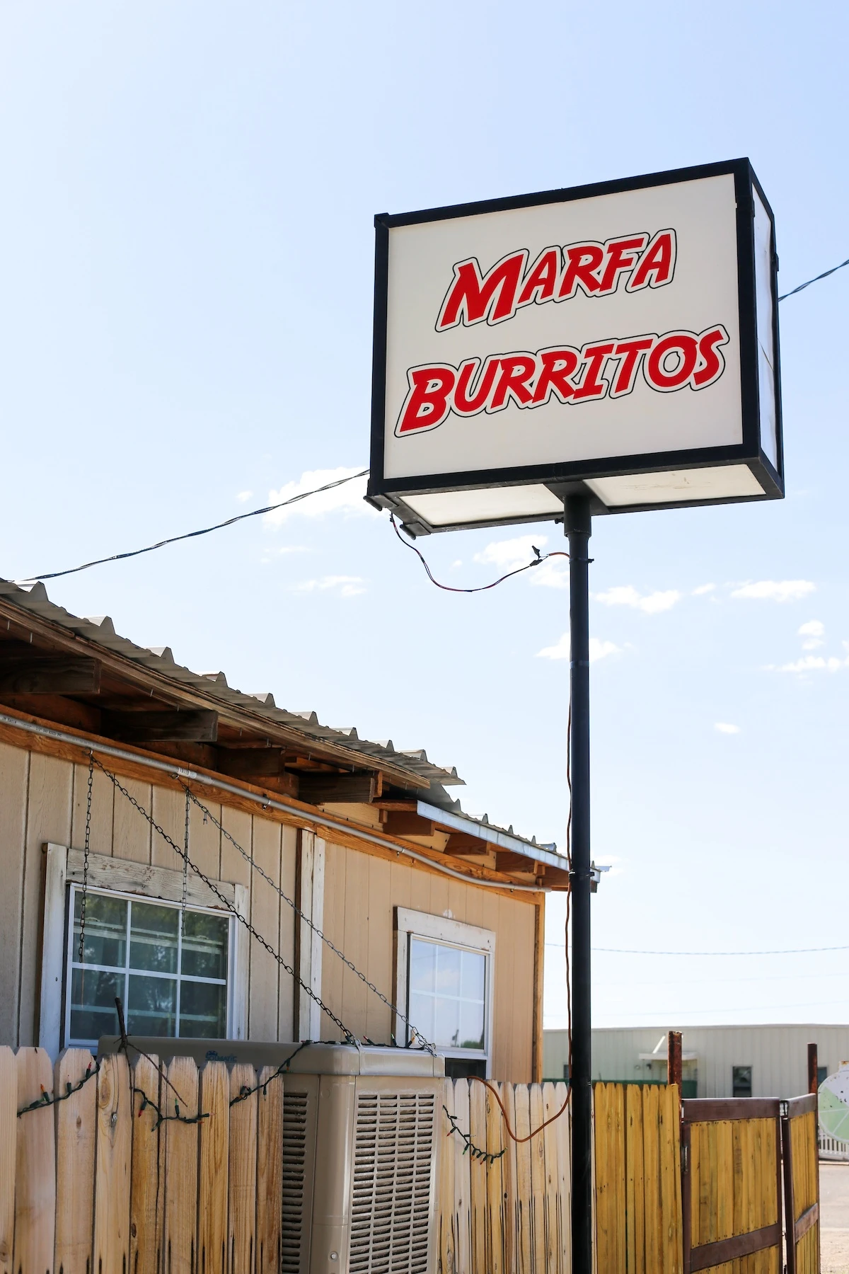 Roadside sign reads "Marfa Burrito" in red letters on white background in front of a beige colored house