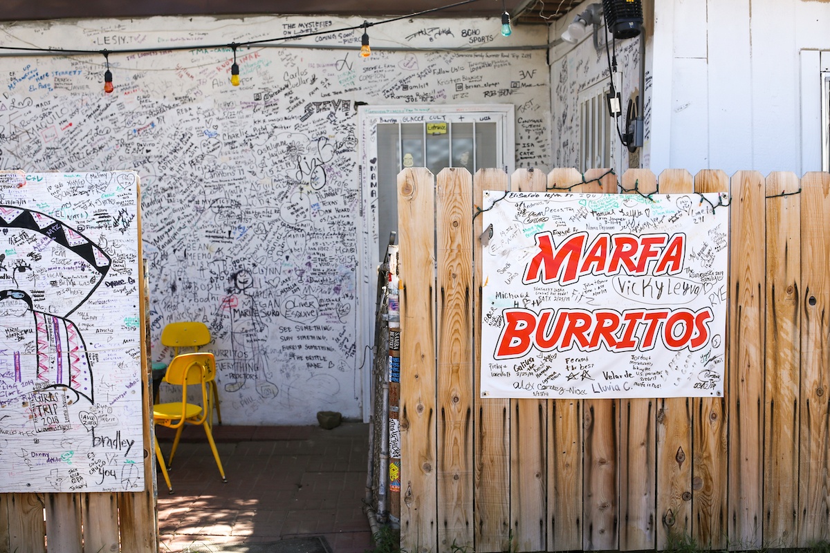 Sign in white with red letters reads "Marfa Burrito" affixed to a wooden fence outside a house. The house and sign are covered with black ink writing and drawings by customers over the years.