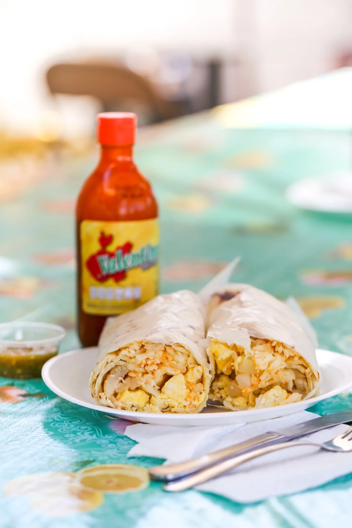 A burrito sliced in half on a white plate atop a table with a bottle of hot sauce and a takeout container of salsa verde behind it