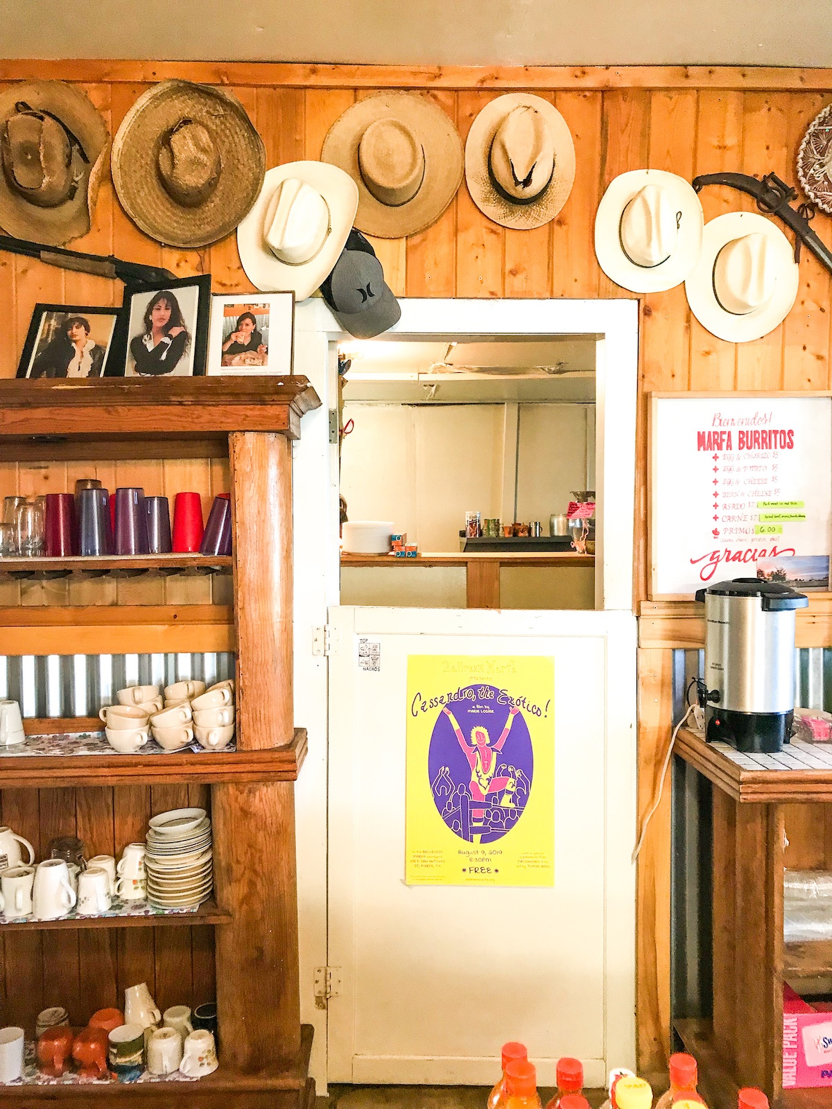 White dutch door in a wood paneled room leads to the kitchen. Cowboy hats hang above the door. To the left of the door are various cups and restaurant items as well as photos of the singer Selena. To the right of the door hangs the menu and an electric hot water container.